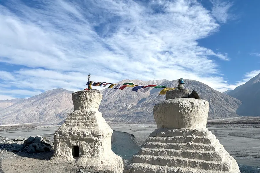 View of ancient white stupas with colorful prayer flags set against barren mountains and a clear blue sky in Ladakh.
