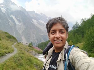 A lady smiling and looking at the camera with the backdrop of mountains .